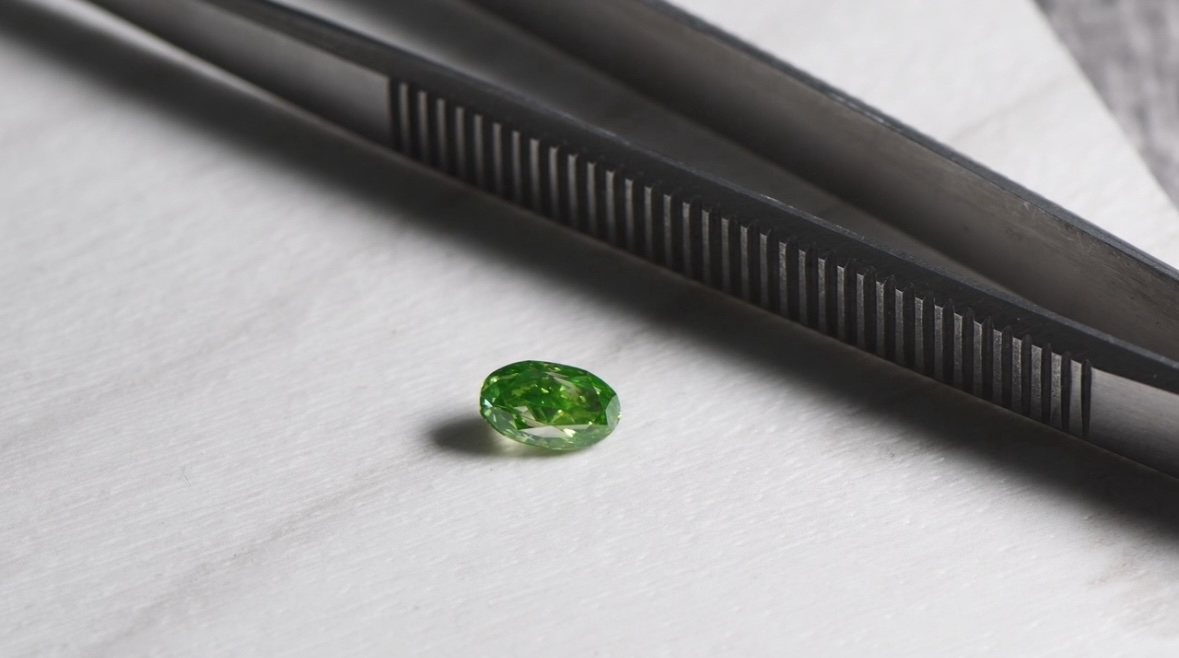 Natürlicher Fancy Green Diamant im Oval Schliff mit Pinzette auf heller Arbeitsunterlage
