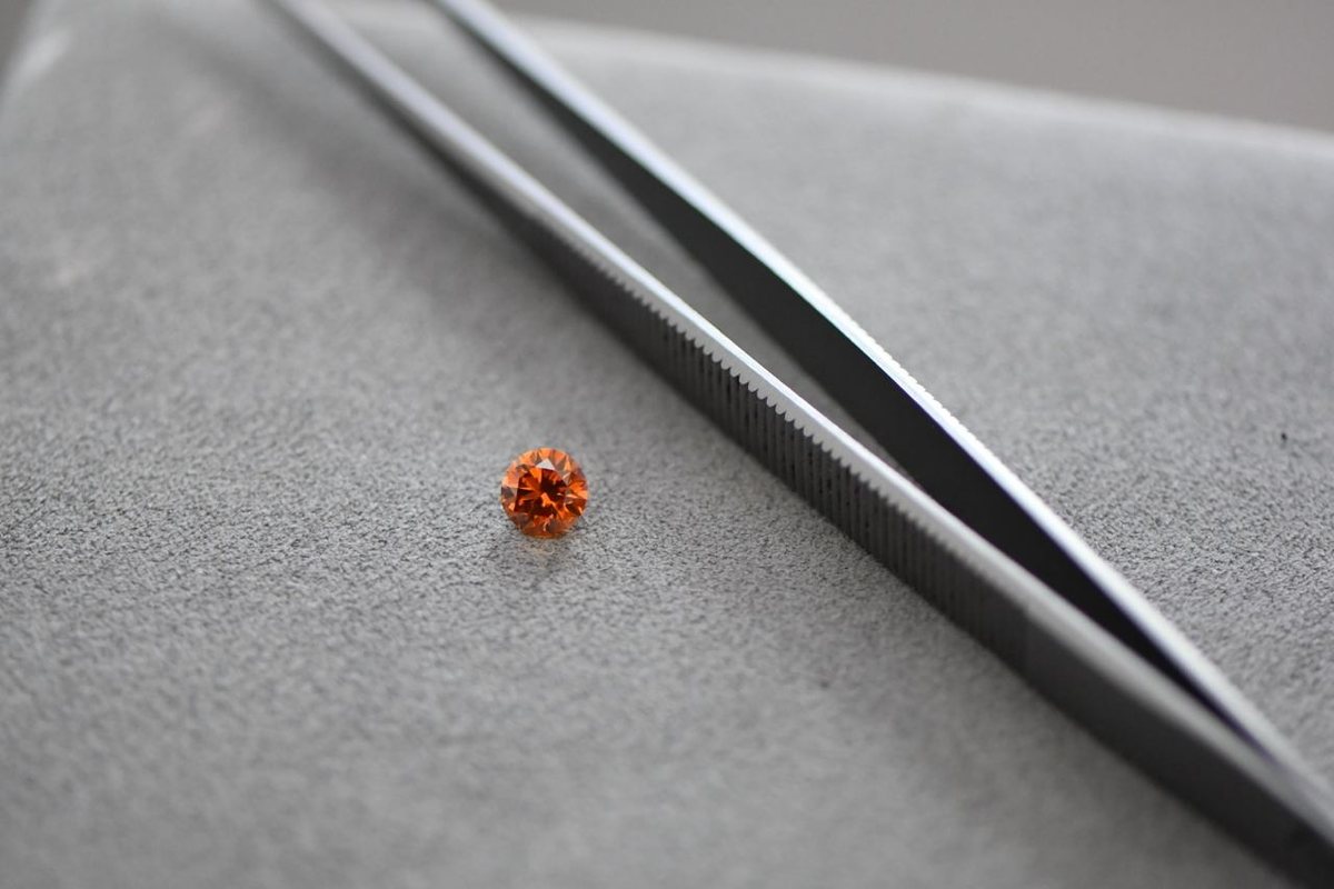Natürlicher Fancy Orange Diamant im Brillant Schliff mit Pinzette auf grauer Arbeitsunterlage